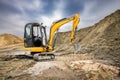 A yellow excavator operates at a construction site, digging into the earth and moving dirt under a cloudy sky in the background Royalty Free Stock Photo