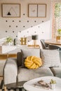 Yellow handmade knot cushion placed on grey couch in white living room interior with crosses and simple posters on the wall