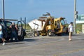 A yellow heavy duty truck hauling a self compacting dumpster along a wooden boardwalk by the beach