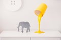 A yellow lamp in the form of a bucket of paint is on a white nightstand. Nearby is a statuette of a gray elephant. In