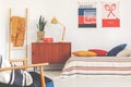 Yellow lamp and plant on wooden cabinet next to bed in sport bedroom interior with posters. Real photo