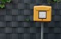 A yellow mailbox stands in front of a gray wall in Germany. A few plants push their leaves through the wall. The emptying times