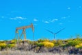 Yellow oil well pump jack sitting on ridge with colorful flowers and folliage with giant industrial windmill peeping over the Royalty Free Stock Photo