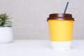 A yellow paper disposable takeout coffee cup with a brown cap, and a straw on white office table with a potted plant Royalty Free Stock Photo