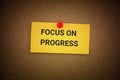 A yellow paper note with the phrase Focus On Progress on it pinned to a cardboard background