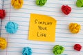 A yellow paper note with the phrase Simplify Your Life on it with some colorful crumpled paper balls around it