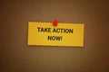 A yellow paper note with the phrase Take Action Now! on it pinned to a cardboard background