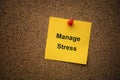 A yellow paper note with the words Manage Stress on it pinned to a cork board