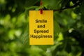 A yellow paper note with the words Smile and Spread Happiness on it attached to a tree branch with a clothes pin