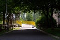 Yellow pedestrian bridge over road turning left from shadow of deciduous trees in park on sunny day