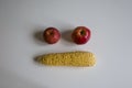 Yellow peeled cob of sweet corn and two red apples formed face emoji two eyes and mouth on white table. Homegrown real organic