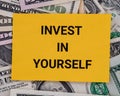 A yellow piece of paper with the phrase Invest in yourself lying on a pile of dollar bills