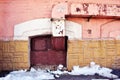 Yellow, pink colors wall with old red boarded up window on the ground level, ventilation pipe, melting snow on asphalt