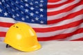 Yellow protective hard hat on light grey table against American flag. Space for text Royalty Free Stock Photo