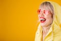 Yellow raincoat. Happy laughing woman on yellow background witn pink heart shaped sunglasses. Bright emotions