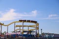 A yellow and red rollercoaster with people riding on the Santa Monica pier with lush green palms trees, blue sky and clouds