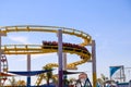 A yellow and red winding rollercoaster surrounded by colorful carnival rides with blue sky and light clouds at Santa Monica Pier