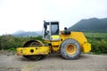 The yellow road roller with a closed cab stands on an uneven road on stones and gravel, side view.