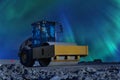 Yellow road roller stands on not ready new road under early night Aurora Borealis, left side view. Northern Lights shine over old