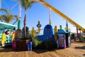 A yellow rollercoaster surrounded by colorful carnival rides and lush green palm trees on a brown wooden pier with clear blue sky