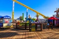 A yellow rollercoaster surrounded by colorful carnival rides and lush green palm trees on a brown wooden pier with clear blue sky
