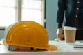 Yellow safety helmet and coffee cup on workplace desk with architect or engineer standing in meeting room Royalty Free Stock Photo