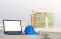 The yellow safety helmet put on the blueprint with laptop has white screen isolated at construction site with crane background Royalty Free Stock Photo