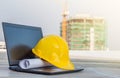 The yellow safety helmet put on the blueprint with laptop has white screen isolated at construction site with crane background