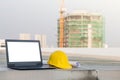 The yellow safety helmet put on the blueprint with laptop has white screen isolated at construction site with crane background Royalty Free Stock Photo