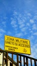 Yellow sign with written in Italian military zone, no access to armed surveillance, on barracks fence