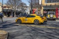 A yellow sports cars stopped at a traffic signal with shops, restaurants and bars, bare trees and people walking on the sidewalk