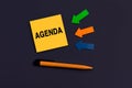 Yellow square sticky note with AGENDA, pen and arrows of paper on dark color background, top view. Business concept. Home Office Royalty Free Stock Photo