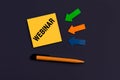 Yellow square sticky note with Webinar, pen and arrows of paper on dark color background, top view. Webinar, online education,