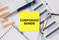 Yellow sticker with text Corporate Bonds lies on financial charts with pen and eyeglass