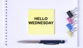 Yellow sticker with text Hello Wednesday lying on a white Notepad with a pen and paper clips