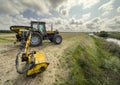 Yellow tractor mowing grass overgrowing the flood bank on the river Royalty Free Stock Photo