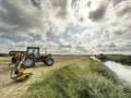 Yellow tractor mowing grass overgrowing the flood bank on the river Royalty Free Stock Photo