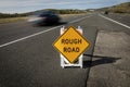 Yellow traffic control sign stating Rough Road on highway shot in late afternoon sun. A car is visible blurred driving past. Royalty Free Stock Photo