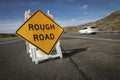 Yellow traffic control sign stating Rough Road on highway shot in late afternoon sun. A car is visible blurred driving past.