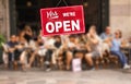 Yes - We Are Open sign in front of blurred people outside a cafe enjoying diner, drinks and coffee between shopping