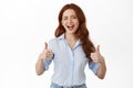 Yes very good. Smiling redhead woman cheering up, winking and showing thumbs up in approval, praise great work, nice job