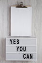 `Yes you can` words on a lightbox, clipboard with blank sheet of paper over white wooden background, top view. Overhead, from Royalty Free Stock Photo