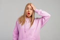 Yikes, oops. Young blonde woman wears violet hoody, stands against grey background surprised with hand on head for mistake,