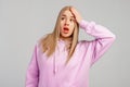Yikes, oops. Young blonde woman wears violet hoody, stands against grey background surprised with hand on head for mistake,