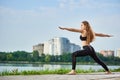 Yoga female standing in a warrior pose, Virabhadrasana, near river in background of urban buildings with copy space Royalty Free Stock Photo