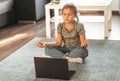 Yoga for kids. Calm little girl in casual wear with closed eyes sitting in lotus pose in front of laptop in living room at home. Royalty Free Stock Photo