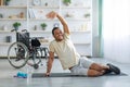 Yoga for paraplegic patients. Disabled black guy stretching on sports mat at home, wheelchair standing nearby