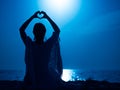 Yoga under full moon over night ocean or sea beach. Young woman's meditation. Girl showing heart shape with hands Royalty Free Stock Photo