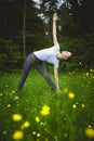 Yoga utthita trikonasana triangle pose by woman white costume on green grass in the park around pine trees