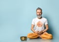 Yogi gray-haired man in dhoti clothes, holding rosary, sitting on floor in lotus pose on blue background. Singing bowl nearby Royalty Free Stock Photo